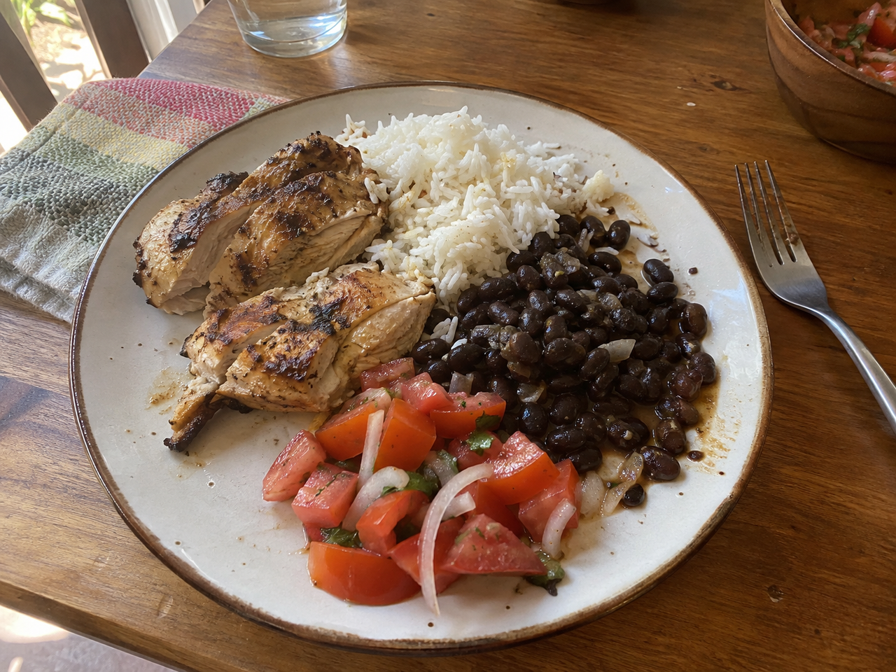 A home lunch of chicken, rice, beans and tomato salad on a kitchen table.